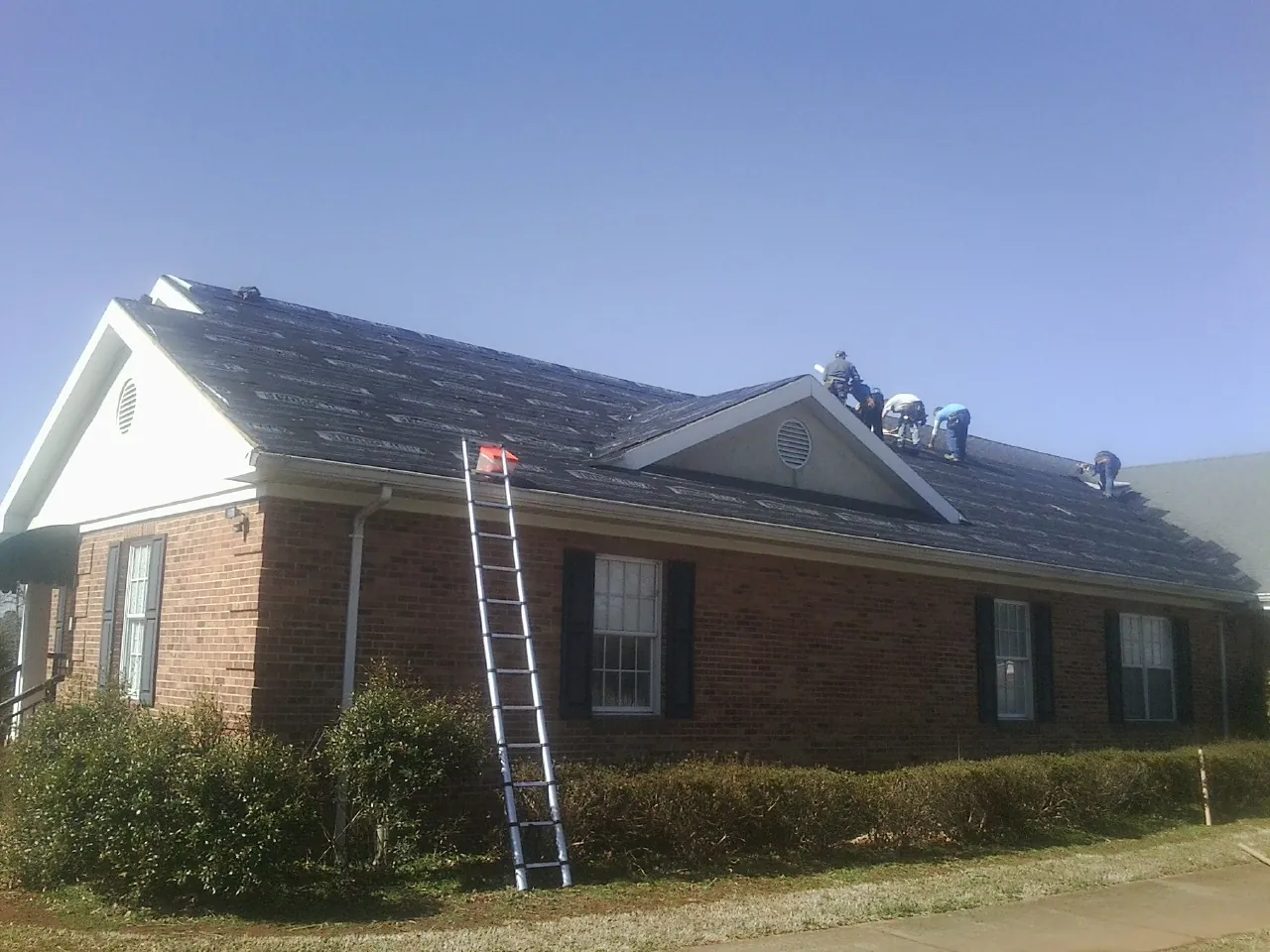 Crew tearing shingles off a brick ranch roof with a ladder set — Emergency Roof Repair work by our crew in Jones