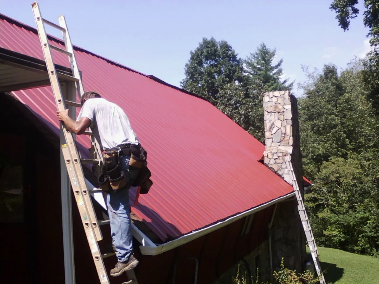 Roofer on a ladder beside a red metal roof and stone chimney — Soffit & Fascia Repair by Roofing Brothers in Jones